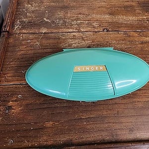 Puede incluir: Una caja de máquina de coser Singer de color azul turquesa con una etiqueta dorada.