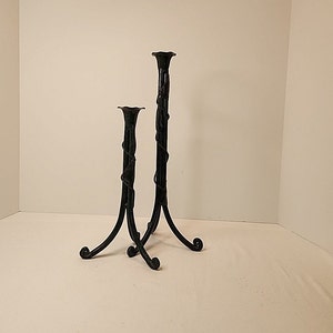 Puede incluir: Dos candelabros de hierro forjado negro. El más alto mide aproximadamente 36 cm de alto y el más bajo unos 25 cm. Ambos tienen un diseño de vid torcida y tapas y bases acampanadas. Los candelabros tienen tres patas curvas.