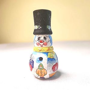 May include: A hand-painted snowman figurine with a black top hat, a yellow scarf, and a painted face. The snowman's body is decorated with winter-themed images, including a wrapped gift and a person in a coat.