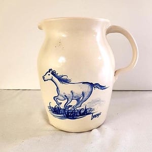 以下が含まれることがあります： クリーム色の陶器のピッチャーで、走る馬の青いイラストが描かれています。 馬はなびくたてがみと尾を持ち、青い草の上に描かれています。 ピッチャーには取っ手と注ぎ口があります。 アーティストのサイン「Laverne」が見えます。