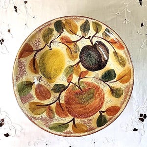 May include: Round ceramic plate with a hand-painted fruit design. The plate features a yellow, orange, and dark plum fruit design with green, orange, and brown leaves. The rim has a speckled brown pattern.