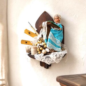 May include: A wall-mounted wooden display shelf featuring a small doll in a bright blue dress with white lace trim. The shelf is decorated with yellow wooden rulers, spools of thread, white lace fabric, and small cream-colored decorative berries.