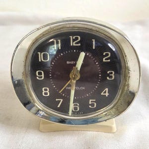 May include: A vintage Westclox Baby Ben alarm clock featuring a round chrome-finished metal frame, a dark brown sunburst dial with white Arabic numerals, and off-white hands. The clock sits on a cream-colored base and displays Made in USA text.