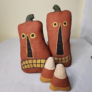 May include: Two orange, rectangular Halloween-themed decorations with black and yellow painted faces and green stems. Two candy corn decorations are in front of the larger decorations. The decorations are on a white surface.