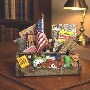 May include: A rustic metal toolbox overflowing with vintage collectibles: an American flag, playing cards, a clock, and small tins. The toolbox rests on a brown leather surface, with a bookshelf and lamp visible in the background.