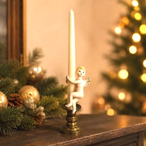 May include: A brass candlestick holder featuring a small ceramic cherub figure with blonde hair and white wings, clutching a tall, slender, off-white taper candle. The cherub is positioned vertically against the candle, which sits in the base.