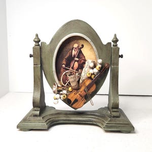Puede incluir: Un marco decorativo ovalado con temática musical en tonos verdes. El marco presenta una imagen central de un violonchelista, con acentos de violín, trompeta y partituras. La base y los soportes también son verdes.