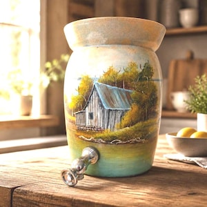 May include: A ceramic beverage dispenser with a hand-painted landscape scene featuring a rustic cabin with a blue roof, surrounded by yellow and green trees. The cylindrical vessel has a wide rim and a silver-toned metal spigot at the base.