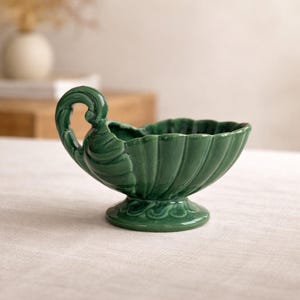 Könnte beinhalten: Eine grüne dekorative Keramikschale mit Griff. Die Schale hat einen gewellten Rand und steht auf einem Sockel. Der Griff ist gebogen und verziert. Die Schale hat eine einfarbige, glänzende grüne Farbe.