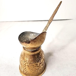 May include: A vintage brass ladle with a decorative floral pattern on the base. The ladle has a small, curved bowl and a long handle with an etched design. The metal has a tarnished, antique appearance.