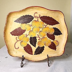 May include: A decorative, vintage tray with a scalloped edge. The tray features a hand-painted design of yellow fruit and dark brown leaves against a cream-colored background. The tray is supported by a decorative metal stand.