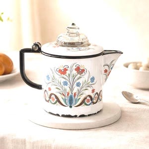 May include: White enamel teapot with a black handle and spout. The teapot features a floral design in red, blue, and brown. It has a clear glass lid and sits on a white circular coaster. A classic kitchenware item.