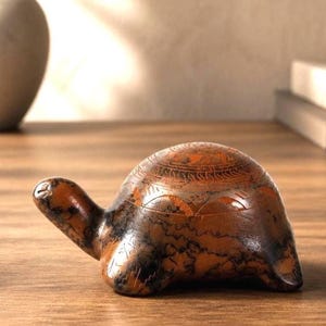 May include: A detailed, brown and black turtle figurine with etched patterns on its shell. The sculpture has a smooth, polished finish and is set on a wooden surface. The turtle's head and legs are extended, giving it a lifelike appearance.