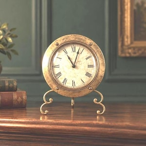 May include: A round, brass-toned table clock with a weathered, off-white face featuring black Roman numerals and two thin black hands. The clock is mounted on a decorative metal stand with two curved, scroll-like feet and a central support.