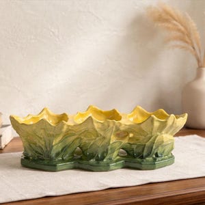 Könnte beinhalten: Ein dekorativer Keramik-Pflanztopf mit Blättermuster. Der Pflanztopf ist hauptsächlich gelb und grün, mit einem gewellten Rand und einem rechteckigen Sockel. Er steht auf einer neutralfarbenen Oberfläche.