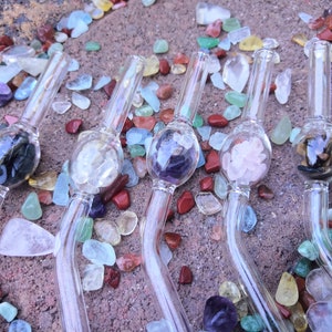 Puede incluir: Cinco pajitas para beber de vidrio con un bulbo redondeado en el extremo. Cada bulbo está lleno de piedras preciosas de diferentes colores. Las pajitas están sobre una superficie cubierta de pequeñas piedras preciosas de colores.