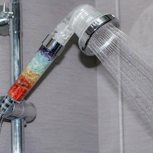 May include: A silver shower head with a clear plastic filter filled with colorful crystals. The shower head is attached to a silver pipe and is spraying water.