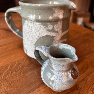May include: A large ceramic pitcher and a smaller matching creamer jug, both featuring a speckled sage green and white glaze with a smooth finish. The smaller creamer has a distinct brown circular stamp on its side near the handle and spout.