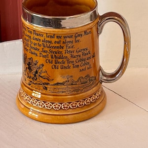 May include: A vintage ceramic beer mug with a silver rim and handle. The mug is tan with a decorative floral pattern at the base and features a poem and illustration. The mug is a classic piece of barware.