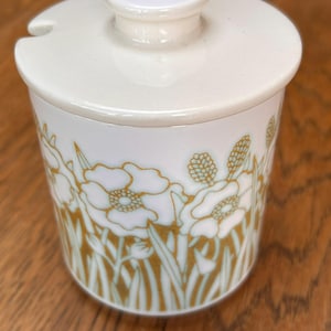 Peut inclure: Un pot en céramique blanche avec couvercle, orné d'un motif floral doré et vert clair sur tout le pourtour. Le couvercle possède un bouton central et une encoche latérale pour une cuillère. Le récipient cylindrique a une finition lisse.