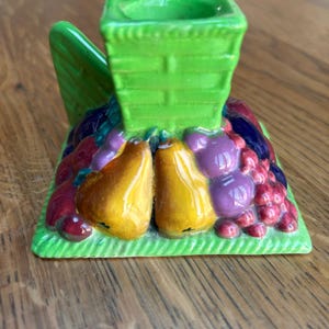 May include: A ceramic candle holder featuring a bright green square base and a central column with a basket-weave texture. The base is decorated with a relief of colourful fruits, including yellow pears, purple grapes, and red berries, with a handle.