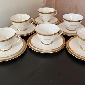 May include: A collection of six white teacups and saucers, each adorned with gold detailing. The teacups feature a gold band on the rim and a gold stripe. The saucers are similarly decorated with gold edging, creating a classic tea set.