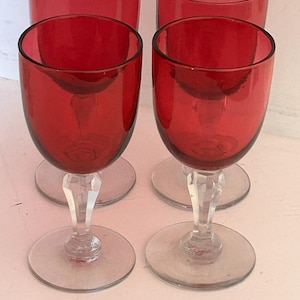May include: Four red wine glasses with clear stems and bases. The glasses have a rounded bowl shape and a vibrant red colour. The stems are faceted, and the bases are circular. The glasses are arranged in a group, suggesting a set for serving.