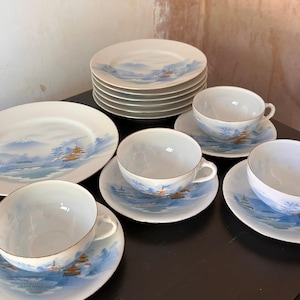 Puede incluir: Una colección de tazas de té, platillos y platos de porcelana vintage. La porcelana blanca presenta un diseño de paisaje azul con montañas y pagodas. Las tazas de té tienen ribete dorado. Los platos están apilados sobre una superficie oscura.
