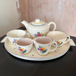 Puede incluir: Juego de té crema con diseños florales. El conjunto incluye una tetera, dos tazas, una jarra de leche y una taza pequeña, todo dispuesto sobre una bandeja ovalada a juego. El estampado floral presenta flores rojas, amarillas y moradas con hojas verdes.