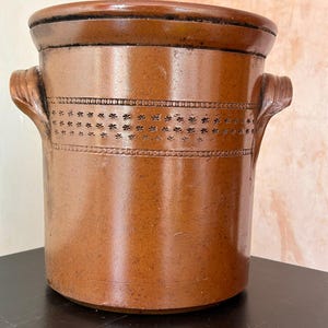 May include: A brown stoneware crock with two handles and decorative floral and dot patterns. The cylindrical container has a wide opening and a dark brown rim. The crock is likely vintage and has a glossy finish.