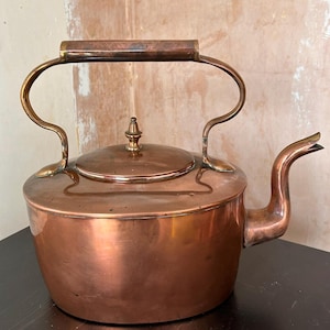 Könnte beinhalten: Ein Kupferkessel mit Messinggriff und Ausguss. Der Kessel hat einen abgerundeten Körper, einen Deckel mit kleinem Knauf und einen gebogenen Ausguss. Der Griff ist gewölbt und mit Messingstützen am Körper befestigt. Der Kessel steht auf einer dunklen Oberfläche.