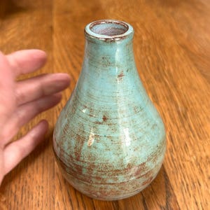 Puede incluir: Un pequeño jarrón de cerámica bulboso con cuello estrecho y base ancha y redondeada. La superficie presenta un esmalte brillante de color turquesa claro con estrías horizontales visibles y detalles rústicos de óxido de hierro rojizo.