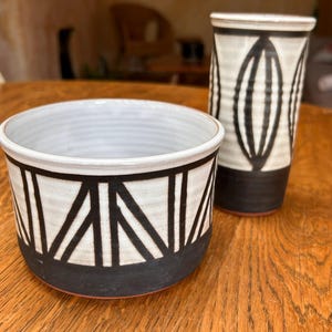 May include: A ceramic bowl and a tall cylindrical vase, both featuring a white glaze with black geometric patterns and a solid black base. The bowl has a wide opening with a triangular design, while the vase displays elongated oval shapes.