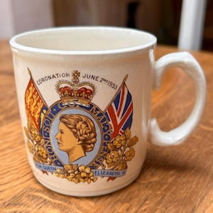 Op de afbeelding: Een crèmekleurige keramische mok met een herdenkingsontwerp voor de kroning van koningin Elizabeth II in 1953. De afbeelding bevat een portret van de koningin, een kroon, twee vlaggen en de tekst Coronation June 2nd 1953 H.M. Queen Elizabeth II.