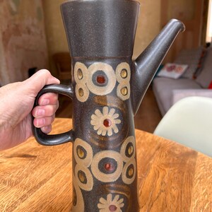 Puede incluir: Una cafetera alta de cerámica marrón oscuro con cuerpo cilíndrico, asa lateral y tapa a juego. El exterior presenta un patrón decorativo de círculos beige y motivos florales estilizados con pequeños centros de color marrón rojizo brillante.