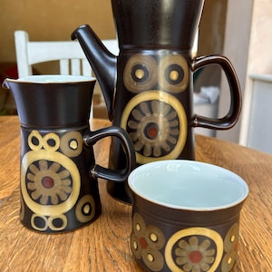 以下が含まれることがあります： 蓋付きの背の高いコーヒーポット、クリーマー、カップで構成されたダークブラウンの陶器製コーヒーセット。各ピースには、ベージュ、オークル、ダークレッドの色合いで円形のパターンが繰り返し描かれており、内側は滑らかな白で仕上げられています。