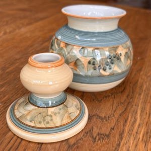 Könnte beinhalten: Eine Keramikvase und ein passender Kerzenhalter mit cremefarbener Basis, horizontalen Streifen in Salbeigrün und einem wiederkehrenden Muster aus stilisierten grünen Blättern und kleinen dunklen Punkten, abgerundet durch orangefarbene Ränder.