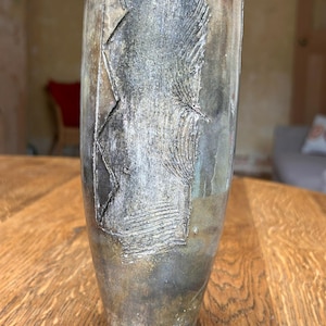 May include: A tall, cylindrical ceramic vase with a mottled, weathered finish in shades of charcoal grey, muted gold, and earthy brown. The surface features a textured, raised relief pattern with vertical lines and geometric zig-zag indentations.