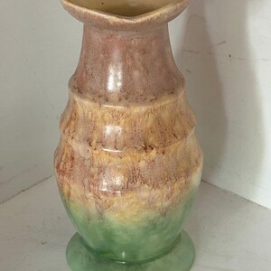Könnte beinhalten: Eine Keramikvase mit herzförmiger Öffnung. Die Vase hat einen Sockel und einen zylindrischen Körper mit geriffelter Textur. Die Farben gehen von Grün am Boden über Gelb und Braun in der Mitte bis hin zu Rosa oben.