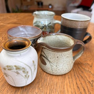 Może przedstawiać: Kolekcja pięciu ceramicznych przedmiotów, w tym dwa małe dzbanki, szeroka miska, wysoki kubek i mały wazon. Przedmioty charakteryzują się różnymi szkliwami w odcieniach ziemi, nakrapianymi teksturami oraz malowanymi motywami roślinnymi w kolorach brązu, kremu i czerni.