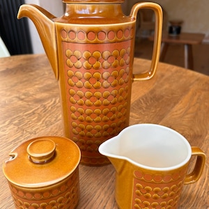 Puede incluir: Una cafetera de cerámica, un azucarero con tapa y una jarra para crema. Cada pieza presenta un esmalte marrón brillante con una banda central de patrones florales repetitivos en naranja y marrón. La cafetera tiene un cuerpo cilíndrico alto.