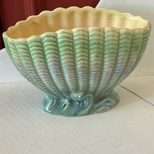 May include: A ceramic seashell-shaped planter with a scalloped rim. The exterior features vertical ridges in shades of green and blue, while the interior is a pale yellow. The planter sits on a small, teal-colored base.