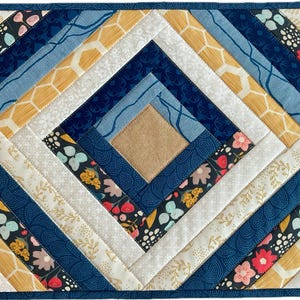 May include: A quilted placemat with a geometric design. The design features a central square of tan fabric surrounded by diamond shapes in various fabrics, including floral, honeycomb, and solid blue patterns. The edges are finished with a dark blue binding.