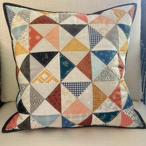 May include: A square throw pillow featuring a patchwork design composed of numerous triangular fabric pieces in various colors including mustard yellow, burnt orange, navy blue, grey, and white, finished with a dark navy blue piped edge.