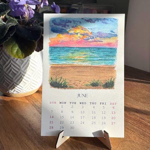 May include: A small desk calendar for June, featuring a watercolor painting of a beach scene with a colorful sunset. The calendar is supported by a wooden stand. A potted plant with purple flowers is in the background.
