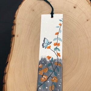 May include: A white bookmark with a blue butterfly and orange flowers painted on it. The bookmark has a green string attached to the top.