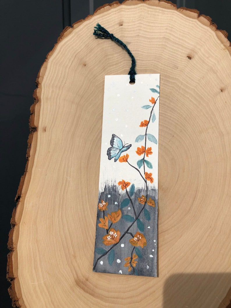 May include: A white bookmark with a blue butterfly and orange flowers painted on it. The bookmark has a green string attached to the top.