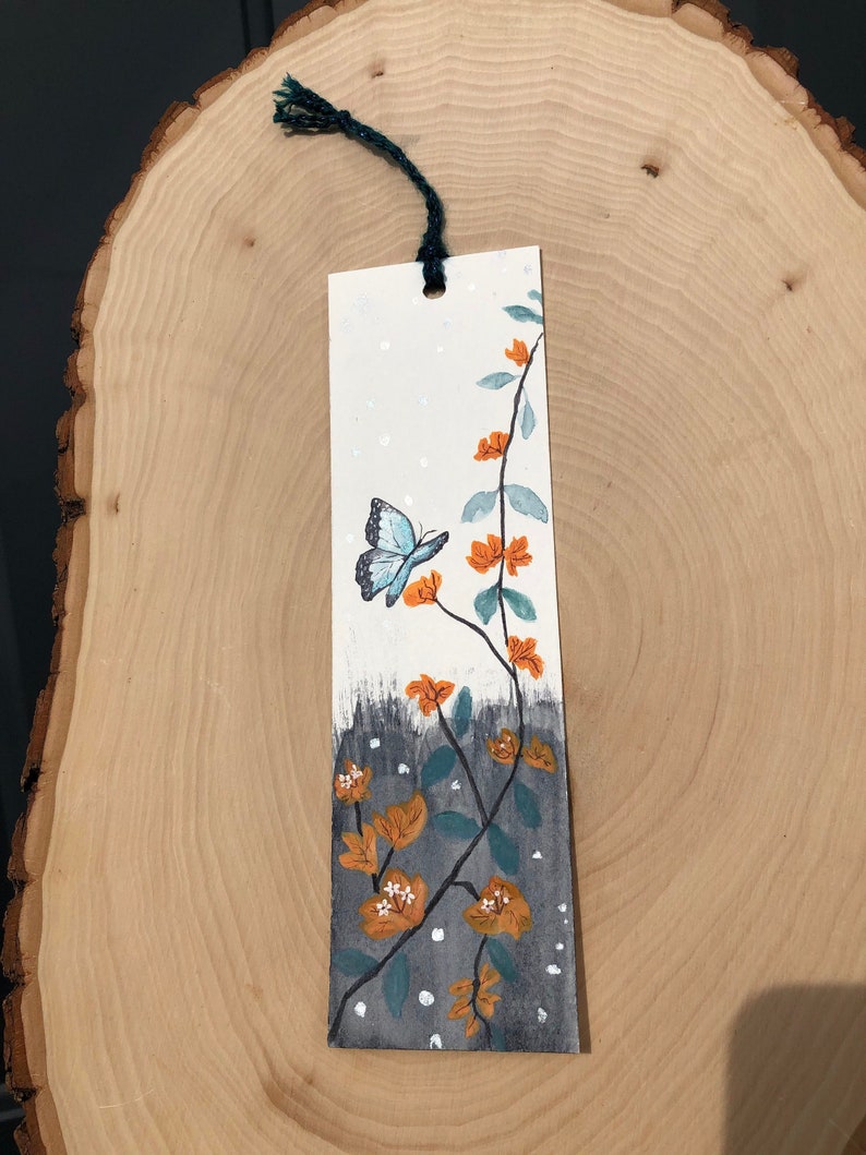 May include: A hand-painted bookmark featuring a blue butterfly flying near orange flowers and green leaves on a white background with gray accents.