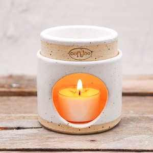 Può includere: Un diffusore di oli essenziali in ceramica bianca con una candela a lumino accesa all'interno. Il diffusore ha un'apertura rotonda e una parte superiore separata. La base e la parte superiore hanno una finitura naturale macchiettata. Il marchio "oo.woo" è stampato sulla parte superiore.