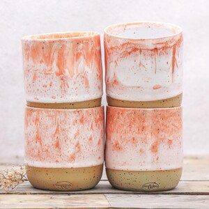 May include: Four ceramic mugs stacked, featuring a speckled, marbled design in coral and white. The mugs have a natural, speckled tan base and a cylindrical shape. The overall aesthetic is modern and handcrafted.